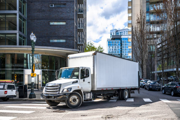 Big rig day cab white semi truck with long box trailer making local commercial delivery at urban city with multilevel residential apartments buildings turning on the city street with crossroad