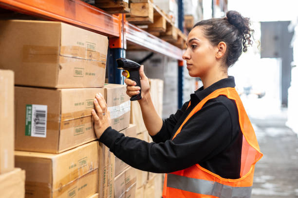 Young female worker scanning boxes with bar code scanner in warehouse shelf. Woman wearing reflective clothing working in modern warehouse.